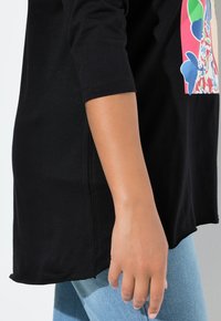Schwarzes Baumwoll-T-Shirt mit 3/4-Ärmeln, das einen bunten Grafikdruck auf der Vorderseite zeigt. Der Saum ist leicht abgerundet und wird mit hellblauen Jeans kombiniert.