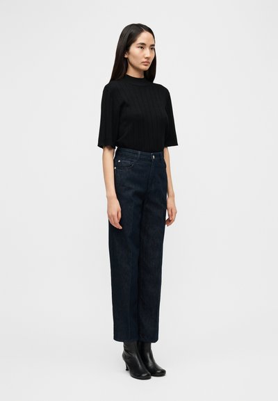 Μαύρο ριμπ κοντομάνικο τοπ σε συνδυασμό με ψηλόμεσα στενά τζιν σκέτης απόχρωσης σκουριάς και μαύρα μποτάκια με τακούνι. Απλό φόντο.