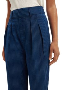 Marineblauwe gepinde op maat gemaakte broek met plooien, voorzien van een brede tailleband en zijzakken, gemaakt van een gladde stof.