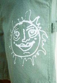 Pantaloni scurți din țesătură verde, cu un grafic alb al unui soare cu față, de formă circulară și raze ondulate, concepuți cu buzunare practice.