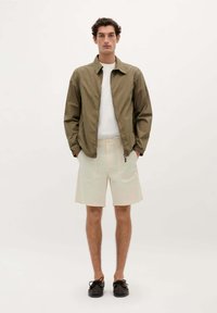 Olivengrøn jakke, hvid t-shirt, beige shorts og sorte sko. Jakken har en lynlåslukning foran og en afslappet pasform.