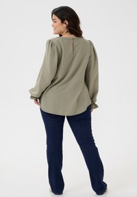 Olive green long-sleeve blouse with puffed shoulders, featuring a back keyhole and a textured fabric, paired with dark blue jeans.