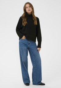 Kvinde med langt brunt hår iført en sort turtleneck sweater, blå brede jeans og sorte sko, stående med den ene hånd i lommen.