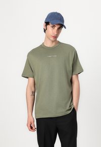 Alyvuogių žaliavos medvilnės marškinėliai su trumpomis rankovėmis ir apvaliu kaklu, su mažuoju baltu "TOMMY JEANS" logotipu. Su derančia mėlyna kepure.