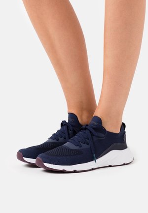 Nogi w granatowych dzianinowych tenisówkach z białymi podeszwami i ciemnofioletowymi spodami, zawiązane na sznurówki, pokazane na białym tle.