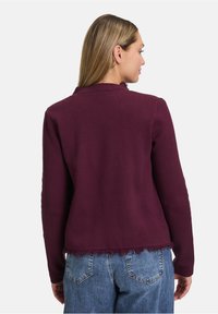 Bordeauxfarbener Pullover mit einem Fransen-Saum, Rundhalsausschnitt und langen Ärmeln, kombiniert mit blauen Denim-Hosen. Weiche Stricktextur, lässige Passform.