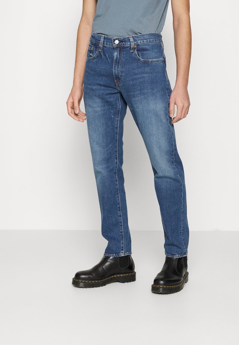 Personne portant un jean bleu moyen coupe droite, associé à des bottines en cuir noir et une chemise bleu-gris atténué.