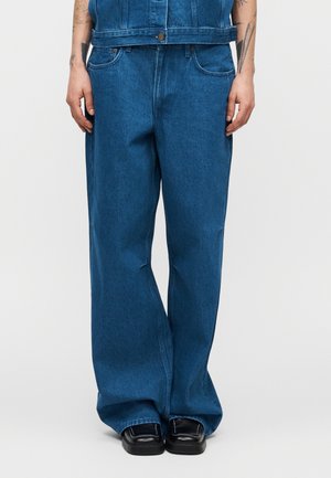 Osoba nosząca luźne, szerokie, niebieskie dżinsy z denimu, czarne buty z kwadratowym noskiem, z widocznymi tatuażami na ramionach, stojąca na tle jasnego, jednolitego tła.