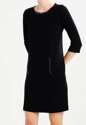 Robe fourreau noire avec des manches trois-quarts, présentant une texture côtelée, un col rond et des accents noirs élégants sur les poches latérales.