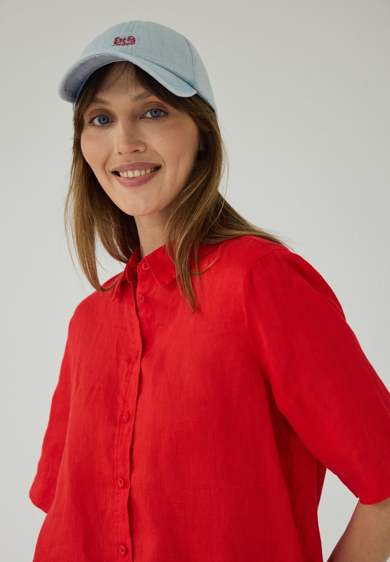 Femme aux longs cheveux châtain clair portant une casquette bleu clair et une chemise rouge vif à boutons, souriante sur un fond uni.