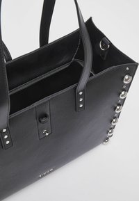Sac fourre-tout en cuir noir texturé avec des clous et des ferrures argentés. Comprend une poche zippée et un intérieur ouvert avec des poignées robustes.