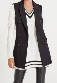 Czarna podwójnie zapinana kamizelka z klapami nałożona na białym swetrze z splotem warkoczowym, posiadającym dekolt w kształcie litery V i czarne paski na brzegu. Czarne skórzane spodnie.