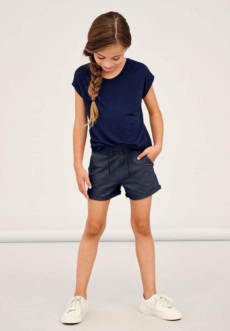 Una camiseta de manga corta de color navy combinada con pantalones cortos grises que cuentan con una cintura elástica y cordón. Terminada con zapatillas blancas.