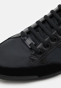 Schwarzer Sportschuh mit einer strukturierten Oberseite aus Stoff und Wildlederakzenten. Mit flachen schwarzen Schnürsenkeln und einem Logo-Tag an der Seite. Glatte Gummisohle.