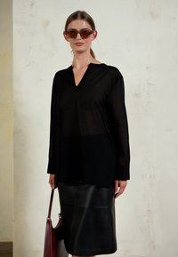 Camisola preta transparente de mangas longas com um design de decote em V, combinada com uma saia lápis de couro preta. A modelo segura uma mala burgundy. Óculos de sol incluídos.