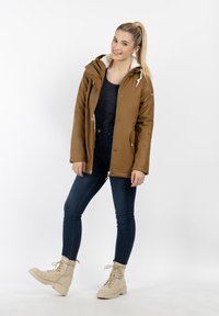 Braune, wasserfeste Jacke mit einer Kapuze aus Kunstfell, Reißverschluss vorne und Druckknöpfen. Tragt über einem schwarzen Oberteil und kombiniert mit blauen Jeans und beigen Stiefeln.