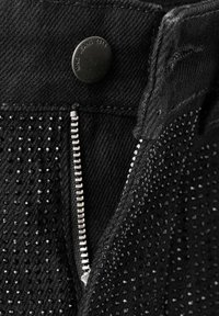 Gros plan sur la fermeture éclair et le bouton d'un jean noir, décorés de petits strass noirs, le bouton gravé "MNG JEANS 1984".