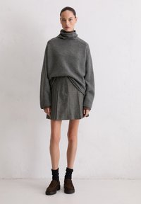 Grijze coltrui, geplooide grijze rok, donkere sokken en bruine suède schoenen. Gladde textuur en casual pasvorm. Effen achtergrond.