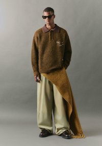 Brauner, gerippter Pullover mit einem dunklen Kragen, beigen, weit geschnittenen Hosen und einem übergroßen braunen Schal, der drapiert ist. Getragen mit dunklen Sonnenbrillen und Schuhen.