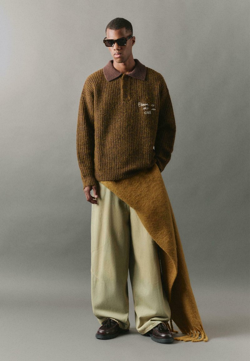 Brauner, gerippter Pullover mit einem dunklen Kragen, beigen, weit geschnittenen Hosen und einem übergroßen braunen Schal, der drapiert ist. Getragen mit dunklen Sonnenbrillen und Schuhen.