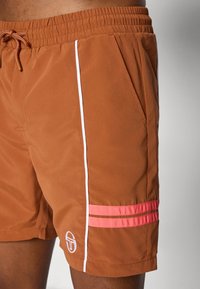Brune badeshorts lavet af let materiale, med hvide sidelister og koralfarvede dekorative striber ved kanten. Inkluderer lommer.