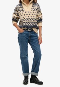 Roomkleurige wollen trui met blauwe geometrische patronen, een halve ritssluiting aan de kraag, gecombineerd met lichtblauwe opgerolde spijkerbroek en zwarte veterlaarzen.