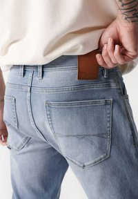 Jeans in denim azzurro chiaro con una vestibilità aderente, dotati di una patch in pelle marrone e due tasche posteriori con dettagli di cucitura discreti.