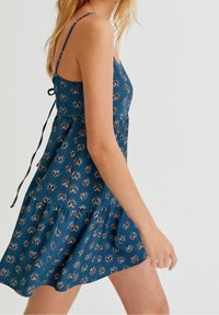 Nainen, joka on pukeutunut hihattomaan siniseen mekkoon, jossa on valkoisia kukkakuoseja, sidottuna takaosasta ohuilla naruilla, kävelee yksinkertaista vaaleaa taustaa vasten.