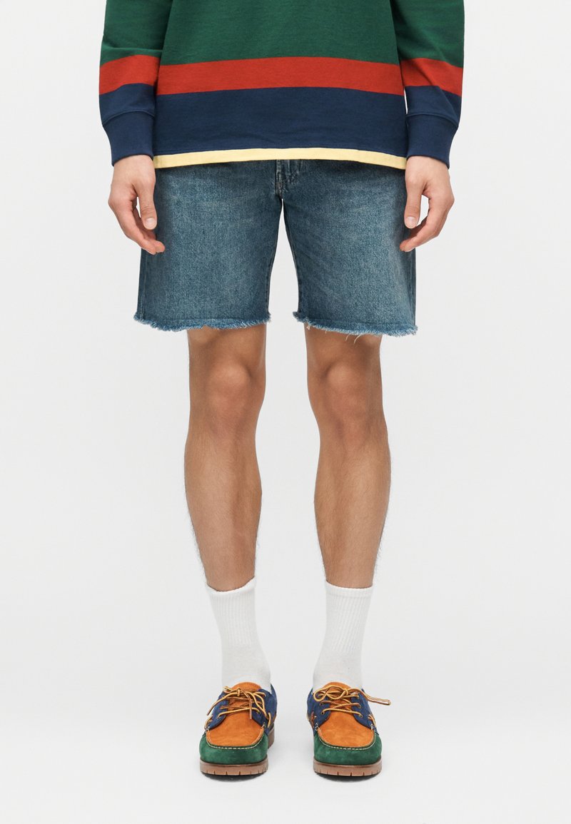 Άνδρας με σκισμένο τζιν σορτς, πράσινο φούτερ με ρίγες σε κόκκινο, ναυτικό και κίτρινο, λευκές κάλτσες και πολύχρωμα παπούτσια τύπου boat shoes.