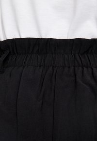 Pantalon en tissu noir taille haute avec une ceinture élastique froncée et une texture plissée, contrastant avec un haut blanc au-dessus.