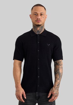 Mann mit Hals- und Armtätowierungen, der ein schwarzes kurzärmeliges, strukturiertes Hemd mit Knöpfen und dunkle Jeans trägt, steht vor einem einfarbigen grauen Hintergrund.