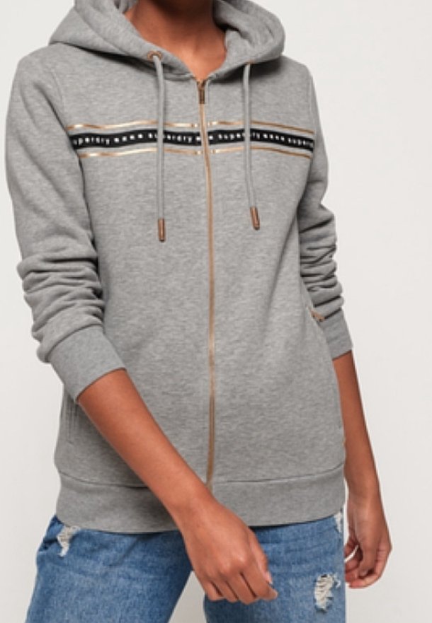 Personne portant un sweat à capuche gris zippé avec des détails rayés et des cordons, associée à un jean bleu déchiré, debout devant un fond uni.