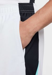 Witte sportshorts met zwarte zijpanelen, met een hand in de zak. De stof heeft een gladde textuur en contrasterende stikdetails.