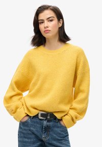 Geel gebreid sweatshirt met een ruime fit, geribbelde textuur, ronde halslijn en verlaagde schouders, gecombineerd met lichtblauwe denim jeans en een zwarte riem.