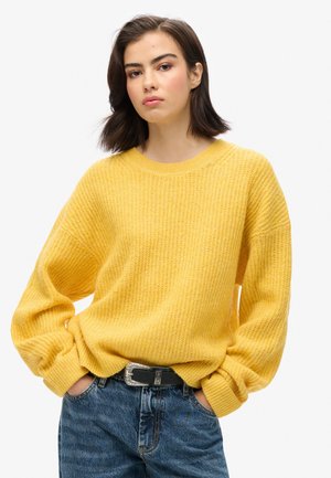 Geel gebreid sweatshirt met een ruime fit, geribbelde textuur, ronde halslijn en verlaagde schouders, gecombineerd met lichtblauwe denim jeans en een zwarte riem.