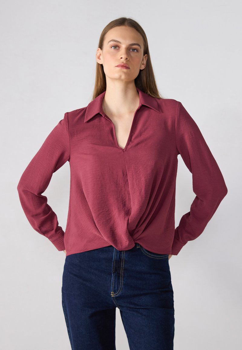 Burgunderfarget langermet skjorte med krage, med en knyttet detalj foran og strukturert stoff. Kombinert med mørkeblå jeans.