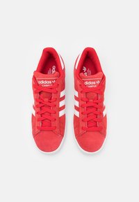 Pár červených tenisek Adidas Campus s bílými pruhy a tkaničkami, pohledem shora na jednoduchém bílém pozadí.