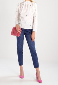 Blouse blanche à manches longues avec broderie d'abeille verte, associée à un jean bleu, des talons aiguilles roses et un sac bandoulière rose en fourrure.