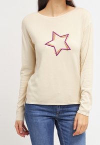 Beige Langarmhemd mit Rundhalsausschnitt und einem lila Sternmotiv auf der Vorderseite. Weicher Stoff und lockerer Schnitt.
