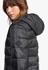 Doudoune noire avec capuche, présentant un design matelassé et une texture douce. Patch logo sur la manche, tissu lisse avec un éclat.