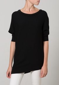 Maglione nero a maglia con scollo ampio adornato da perline nere. Ha maniche corte e l'orlo posteriore leggermente più lungo. Indossato su pantaloni bianchi.