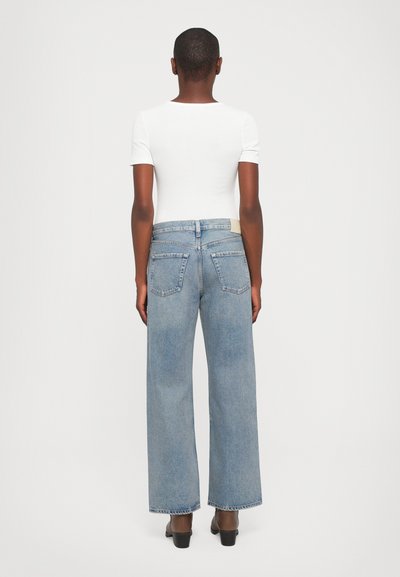 Light blue wide-leg jeans in denim, featuring five pockets and a small beige label on the back waist. Worn with a white fitted top.