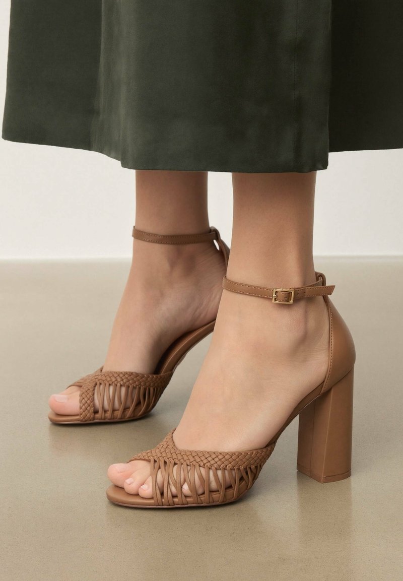 Pieds portant des sandales à talons hauts couleur fauve avec des brides tissées à l'avant et une boucle à la cheville, associées à un pantalon court vert foncé.