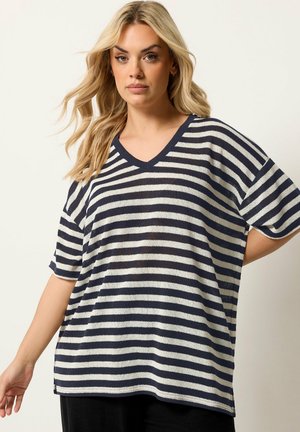 Blonde Frau trägt ein lockeres, marineblau-weiß gestreiftes V-Ausschnitt-Top mit kurzen Ärmeln und posiert vor einem einfarbigen hellen Hintergrund.