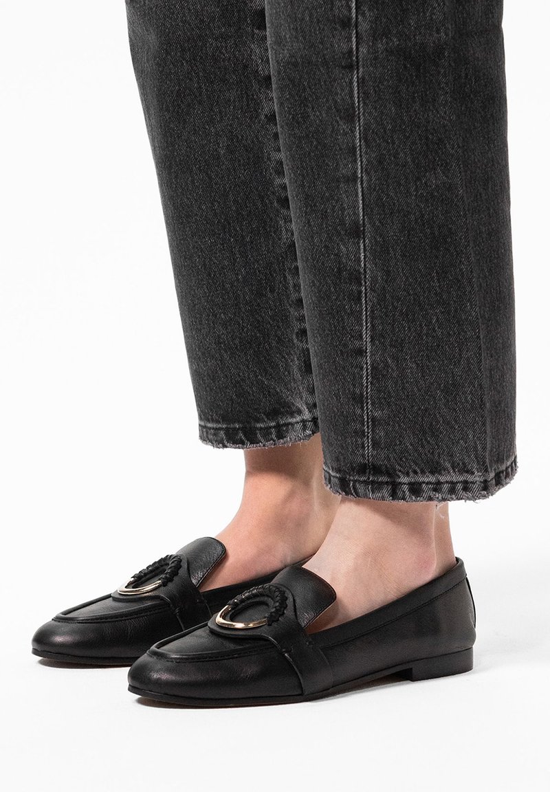 Zwarte leren loafers met een afgeronde neus, voorzien van een decoratieve gevlochten accent en gouden hardware. Plat, laag profiel met open zijkanten.