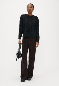 Μαύρο ζακέτα πλεκτού τύπου κασμίρι με κλείσιμο με κουμπιά και κορδόνι στη μέση, συνδυασμένο με καφέ παντελόνι καμπάνα από κορδόνι και μαύρα παπούτσια με πλατφόρμα.