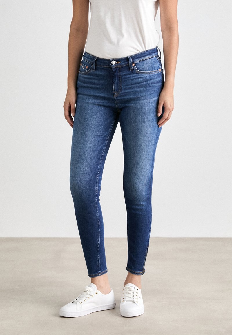 Vaqueros ajustados de color azul con cierre de botón, trabillas para cinturón y un leve desgaste. Combinados con una camiseta blanca y zapatillas blancas.