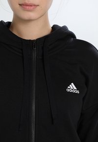 Sweat à capuche noir zippé en tissu doux, doté d'une capuche, d'une fermeture éclair à l'avant et du logo Adidas blanc sur la poitrine gauche.