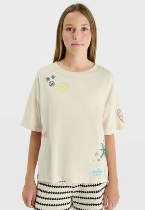 Jeune femme portant un t-shirt ample crème avec de petites imprimés colorés de fleurs, de soleil, de cœur et de palmiers, associé à un short à motifs noirs et blancs.
