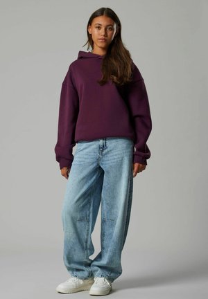 Lila Kapuzenpullover mit überschnittenen Schultern, kombiniert mit hellblauen weiten Jeans und weißen Sneakers. Glatte Stoffoberfläche, lässiges Design.
